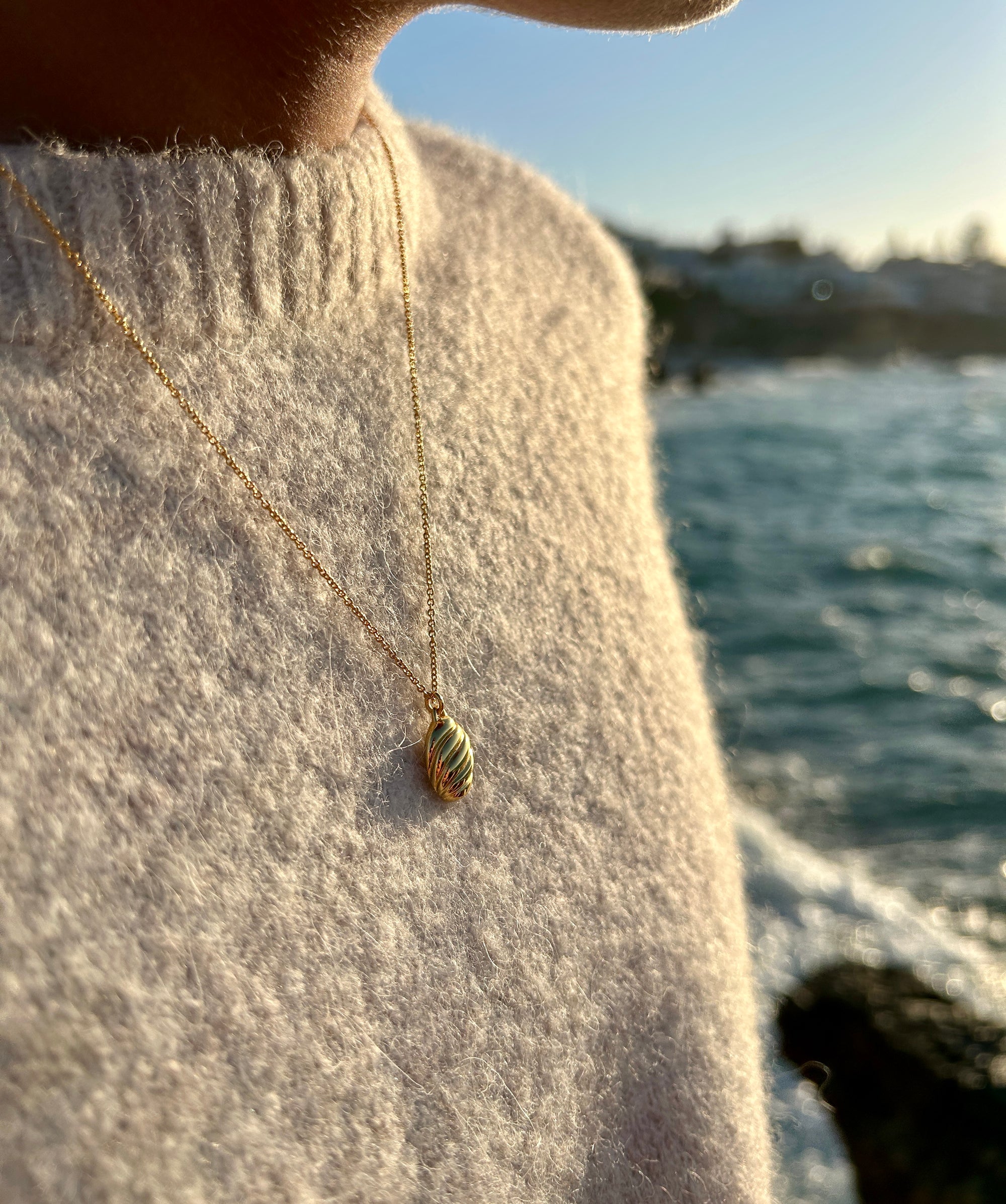 Guldhalsband dam med detaljrik design hängandes runt en mänsklig hals liggande mot bröstkorgen med en bakgrund av blått hav.
