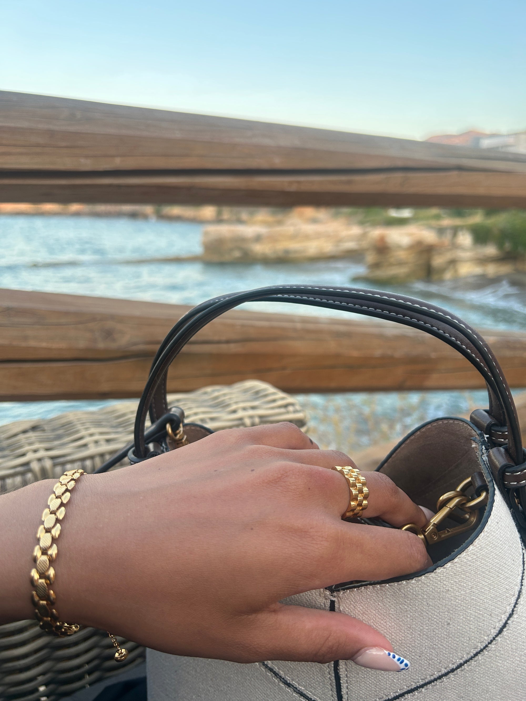 Armband dam i guld med detaljrik och elegant design runt en mänsklig handled som sträcker sig mot öppningen av en väska med en bakgrund av två trä stänger 